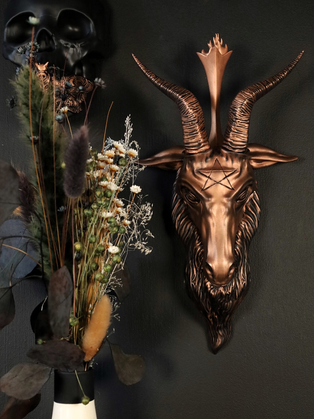 Bronze Baphomet goat head wall decoration on a dark background with dried plants.