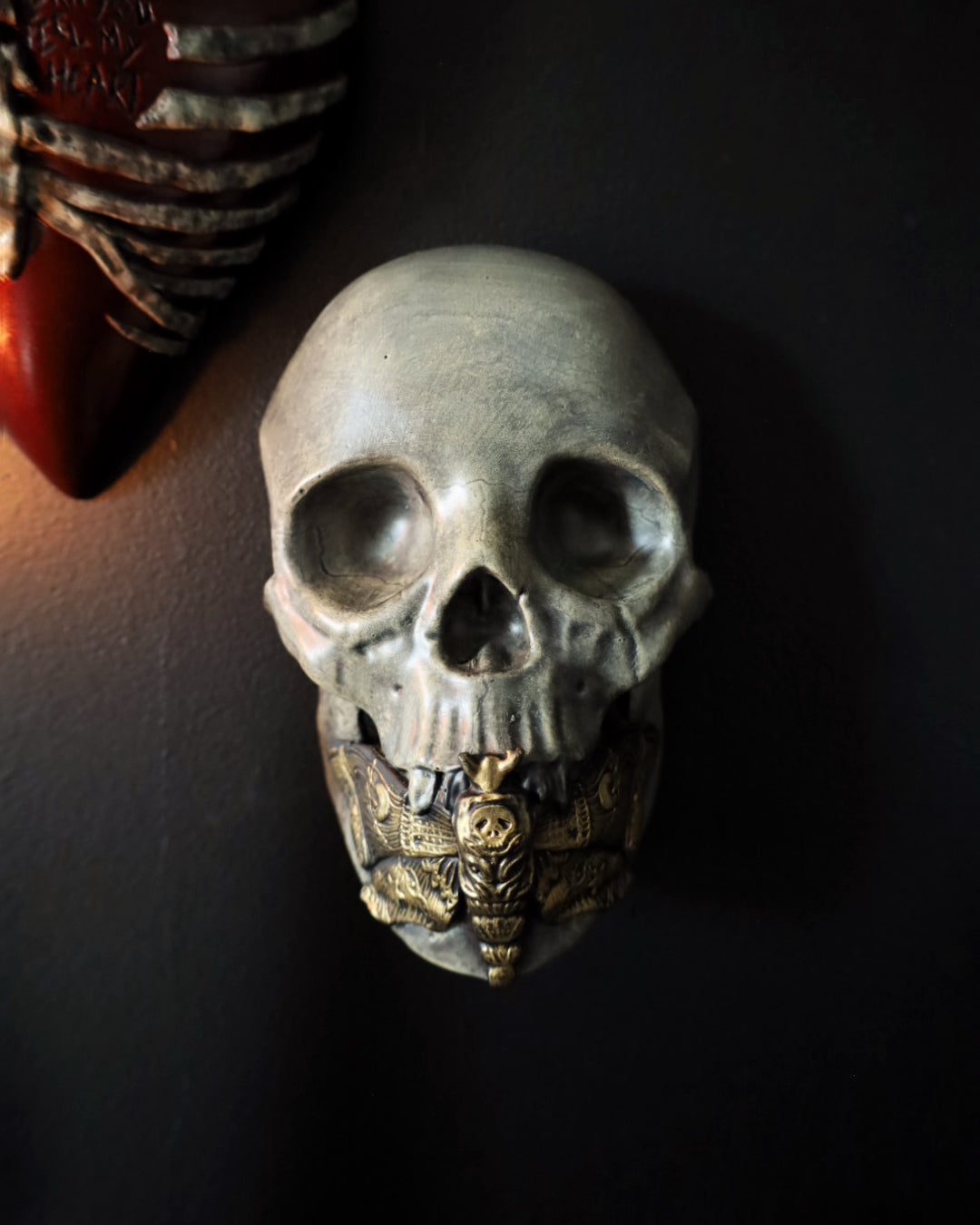 A decorative gothic skull wall hanging with a weathered grey finish and gold toned deaths-head hawkmoth detail at the jaw