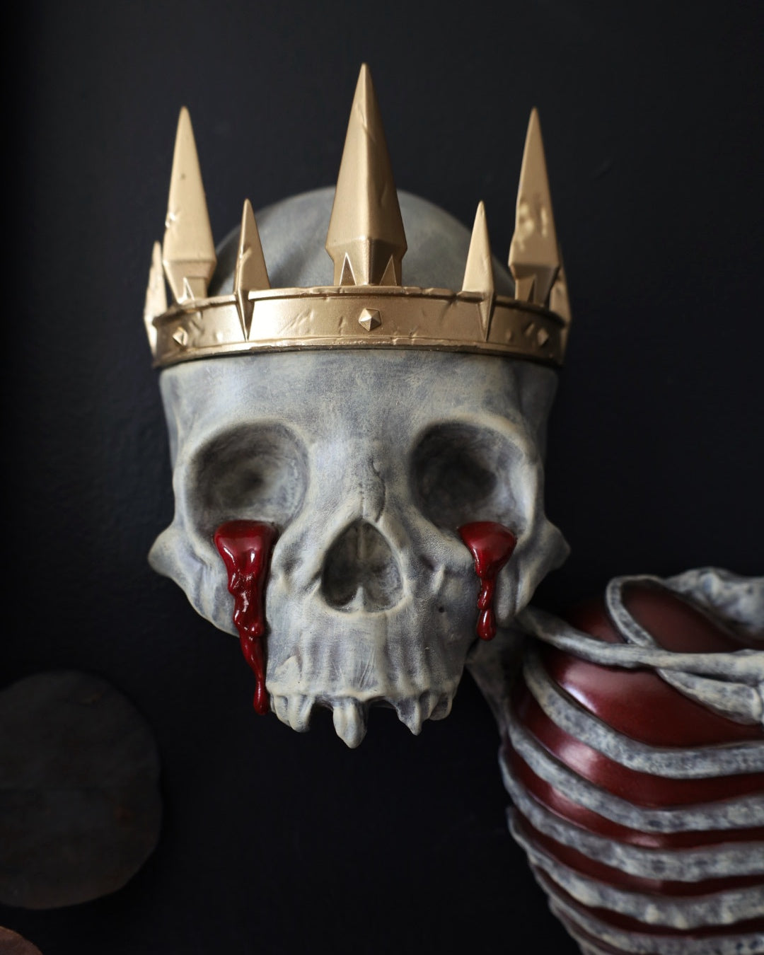 A decorative gothic skull sculpture wearing a gold crown, with red blood-like tears from the eyes. A striking piece for any gothic gallery wall