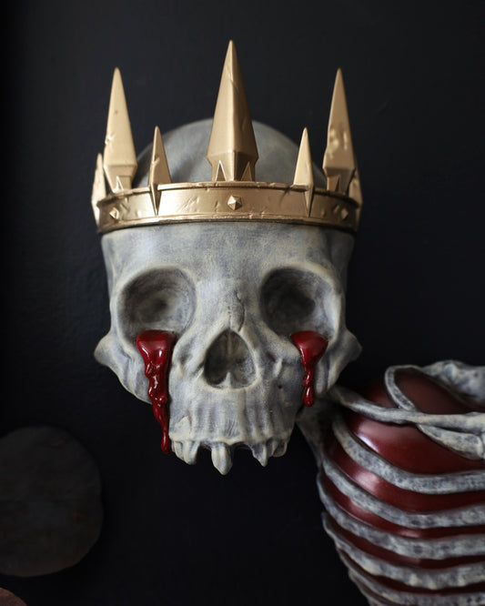 A decorative gothic skull sculpture wearing a gold crown, with red blood-like tears from the eyes. A striking piece for any gothic gallery wall