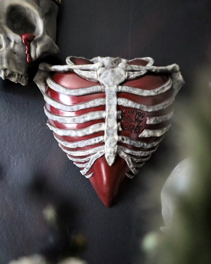 A gothic style wall plaque shaped like a ribbed human chest with a glossy red heart and weathered bones