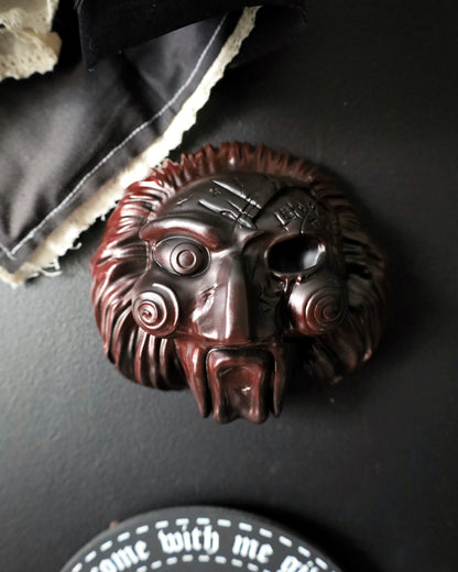 A dark red/black fade jigsaw character with spiral eyes and a wide open mouth, from the Saw movies