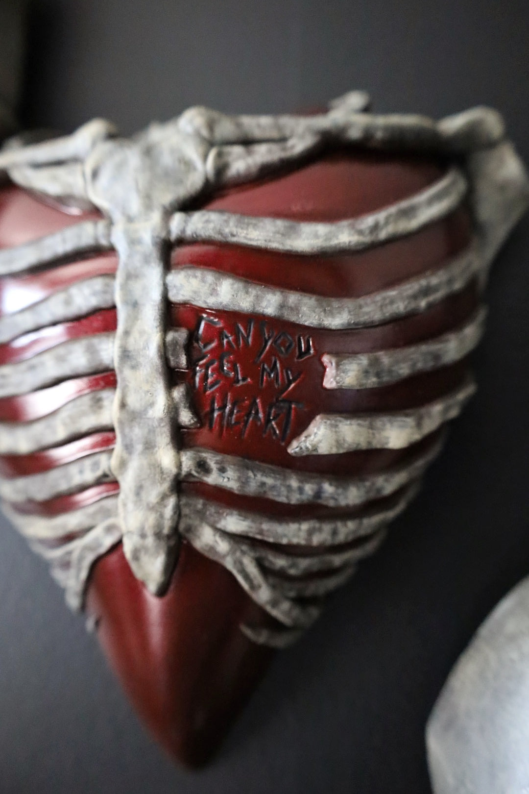A close-up of a red rib cage sculpture wall hanging, featuring the engraved text 'CAN YOU FEEL MY HEART' on the front, set against a dark background.