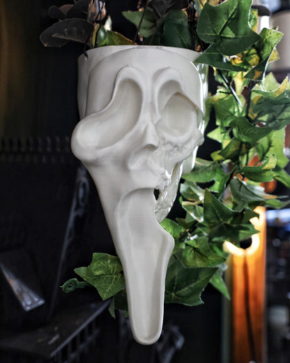 A white ghostface hanging planter in the shape of a skull with overgrown green plants.