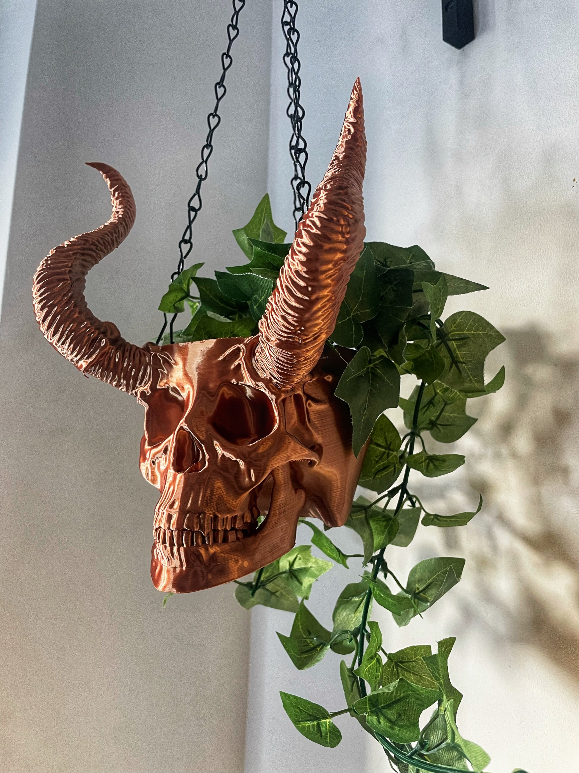 A copper horned skull hanging planter with green plants growing out of it, suspended by a chain.