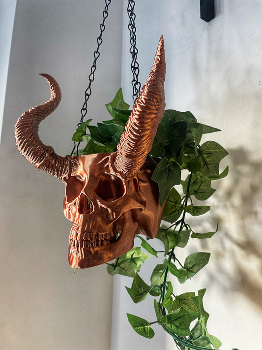 A brown horned skull hanging planter with green plants growing out of it, suspended by a chain.