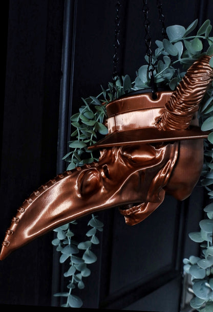 A copper colour hanging planter designed in the shape of a plague doctor's mask, adorned with greenery. Gothic home decor or a great gothic organiser.