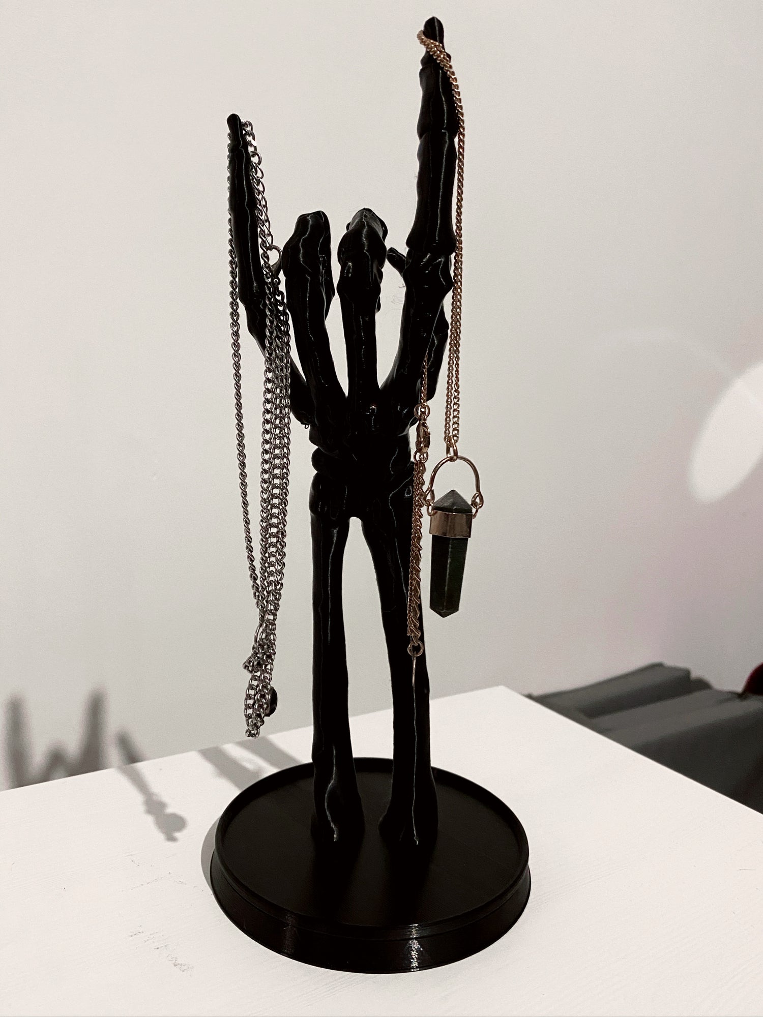A black skeleton hand jewellery stand with a necklace and a ring on it, placed on a white surface.
