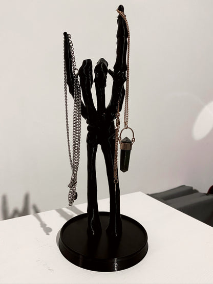 A black skeleton hand jewellery stand with a necklace and a ring on it, placed on a white surface.