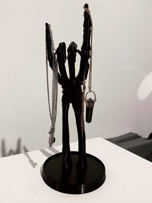 A black skeleton hand jewellery stand with a necklace and a ring on it, placed on a white surface.